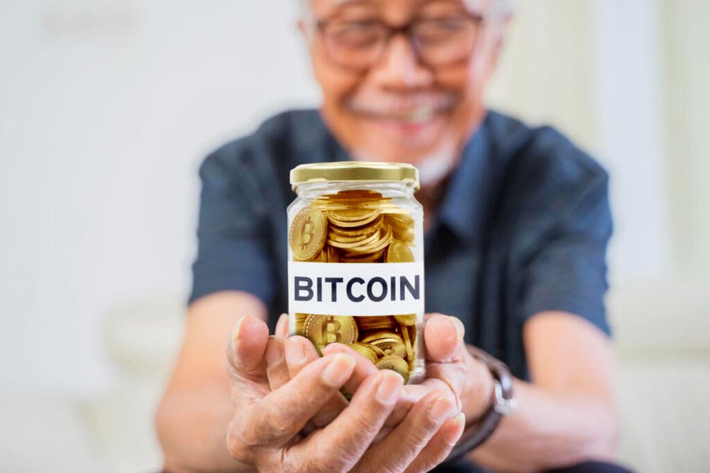 An elderly man is smiling and holding a glass jar filled with golden Bitcoin coins, with a label on the jar that says "BITCOIN".