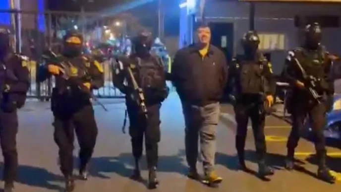 A man in a polo shirt and jeans is being escorted by a group of heavily armed and masked police officers at night