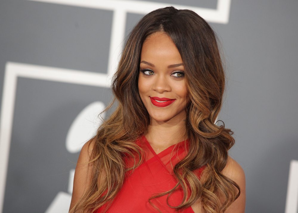 Rihanna with long, wavy brown hair, wearing a red dress, smiles at the camera