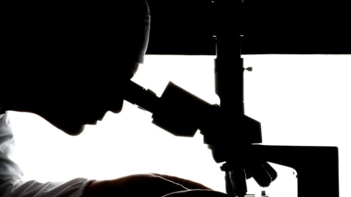 The silhouette of a person in a lab coat looks through a microscope against a bright white background