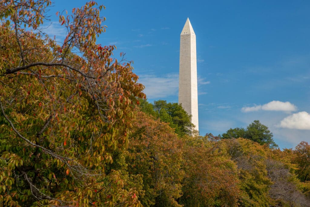 The top of the Washington Monument, a white obelisk, is visible above a hill covered in colorful autumn foliage