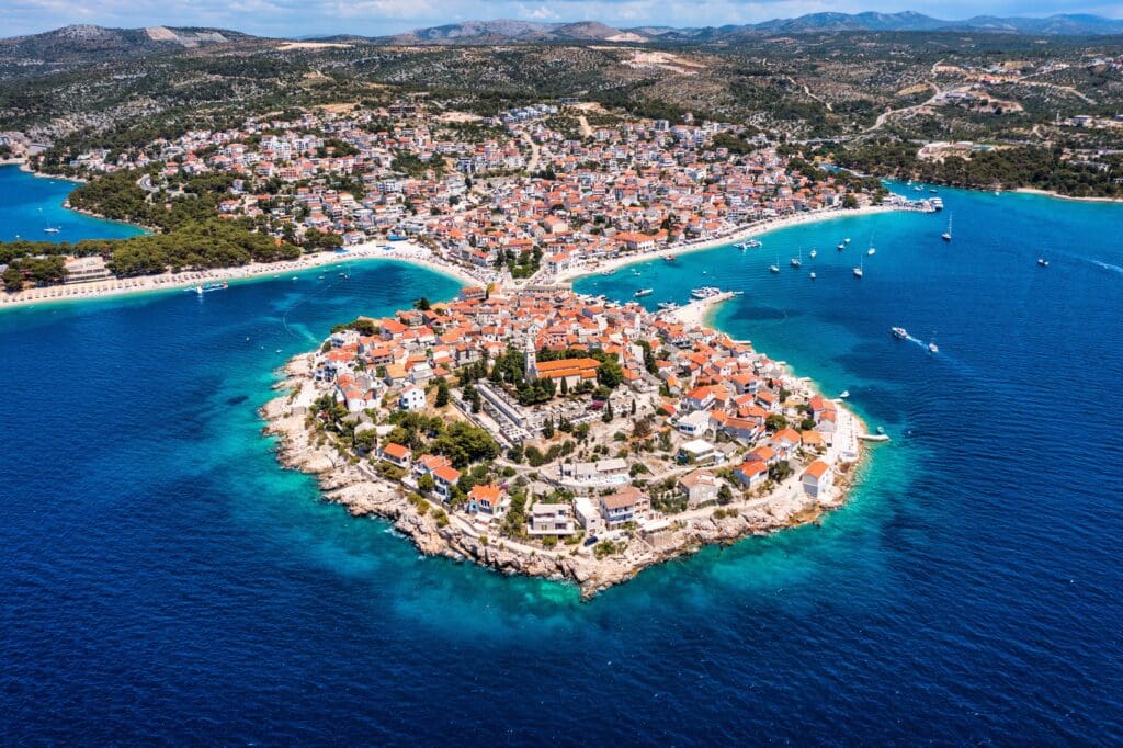High aerial view of Primosten, a historic Croatian town built on a small, round islet in the Adriatic Sea, connected to the mainland by a causeway.
