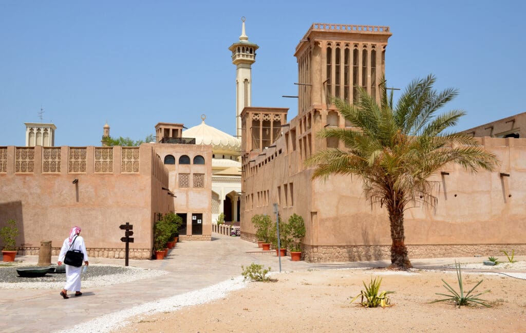 A person in traditional attire walks through the sandy courtyard of Al Bastakiya in old Dubai, featuring sand-colored buildings and a palm tree.