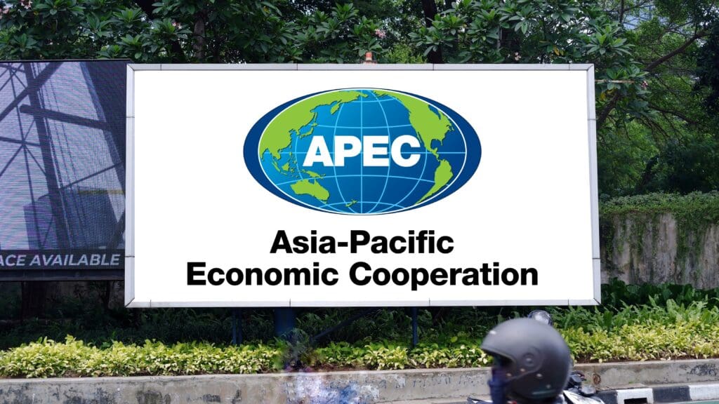 The APEC (Asia-Pacific Economic Cooperation) logo and name displayed on a large roadside billboard in front of lush greenery.