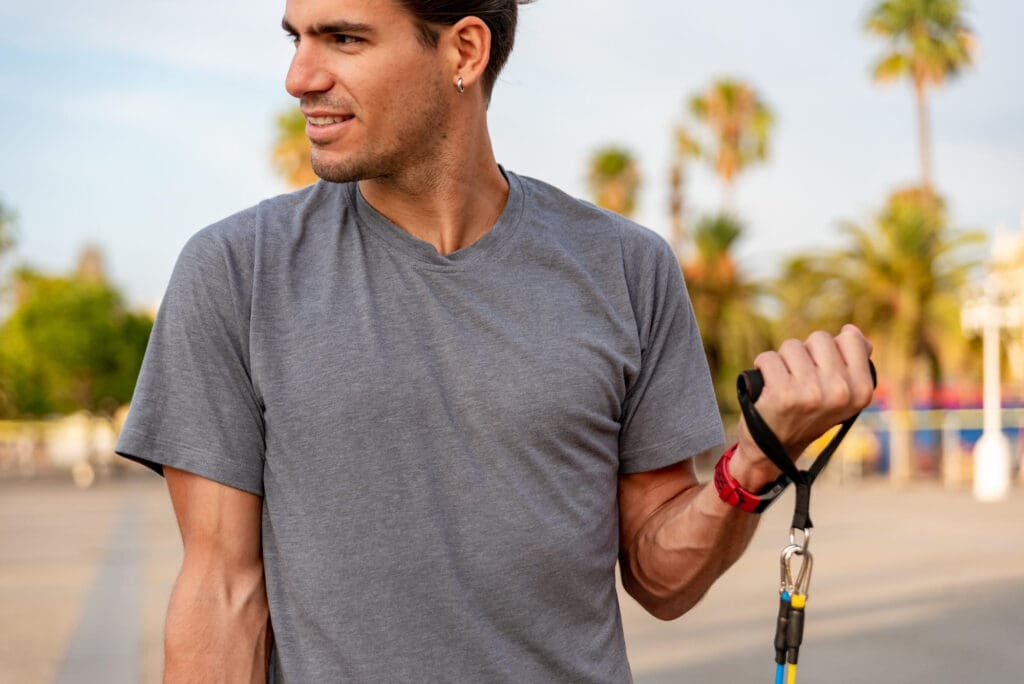 Man in a gray V-neck athletic t-shirt holds resistance bands, ready to exercise in a sunny, palm-tree-lined park.