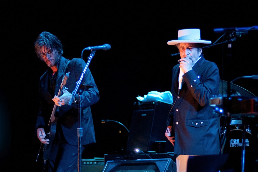 Singer Bob Dylan in a white hat and black suit stands at a microphone next to his guitarist who plays a solid-body electric guitar.