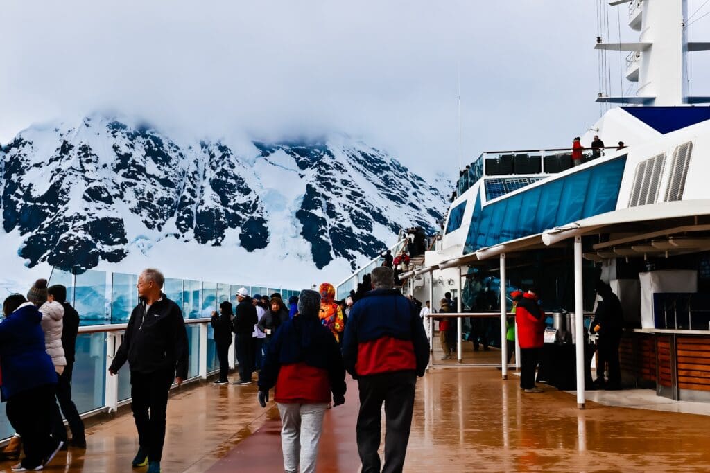 Cruise ship passengers walking on a damp deck, gazing at a large, cloud-shrouded, snow-covered mountain in Antarctica