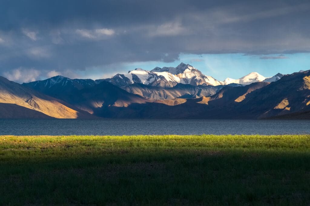 A dramatic landscape of Tso Moriri Lake in Ladakh, India, with sunlit, snow-capped mountains.