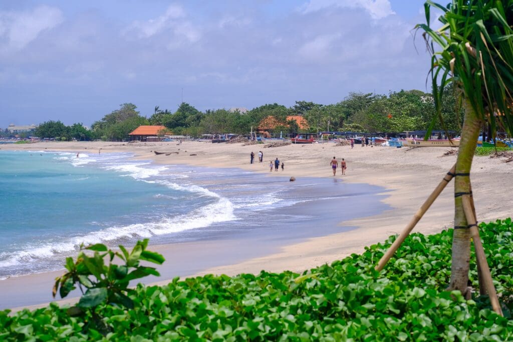 A view of Jerman Beach in Kuta, Bali, showing people walking on the sand, turquoise water, and boats.
