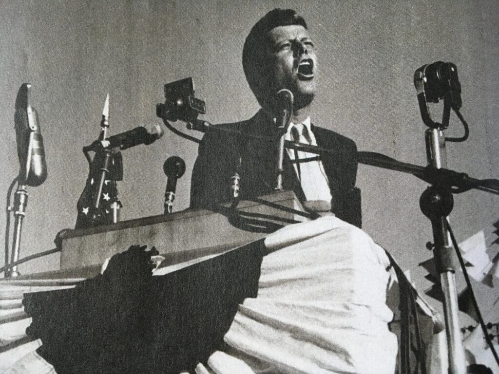 Black and white photo of John F Kennedy passionately addressing a crowd from a podium, with his mouth open and multiple microphones surrounding him.