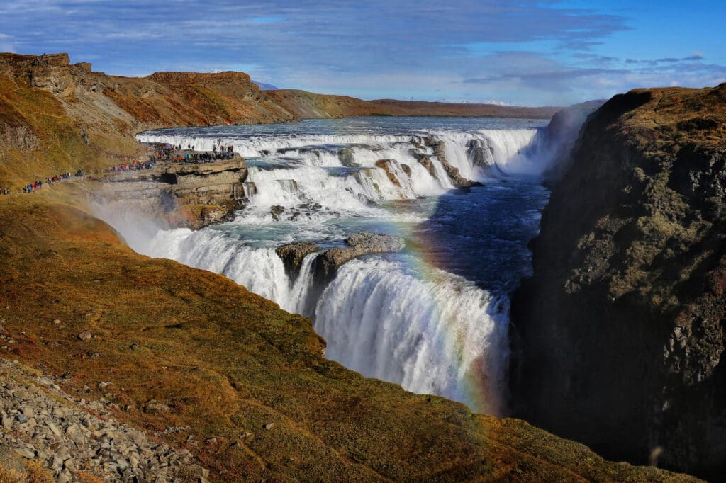 Wide, multi-tiered waterfall cascading into a deep canyon with a rainbow arching through the spray, surrounded by brown and red scrubland.