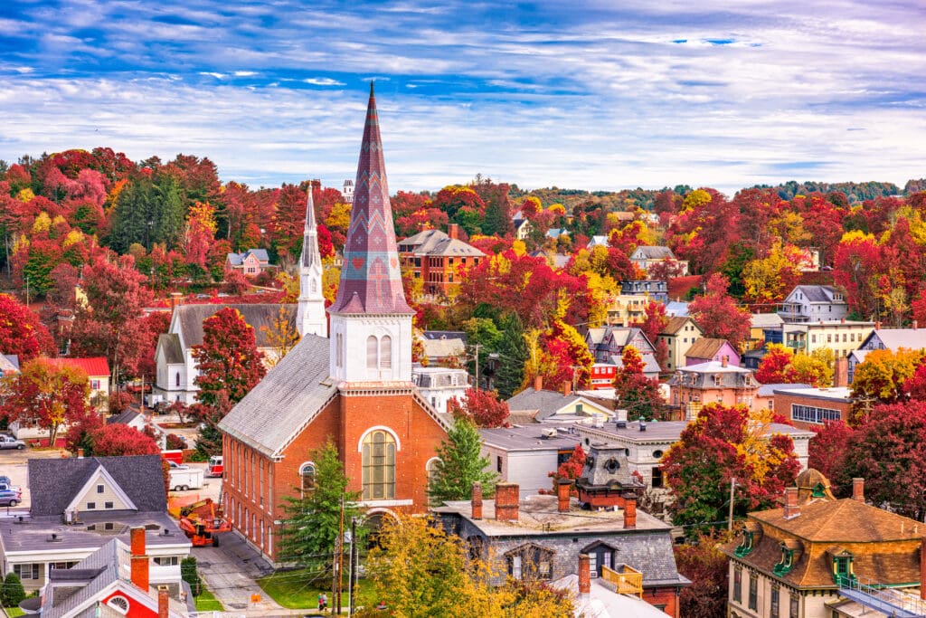 An elevated view of Montpelier, Vermont, showing a town skyline dominated by a brick church spire surrounded by vibrant autumn foliage.