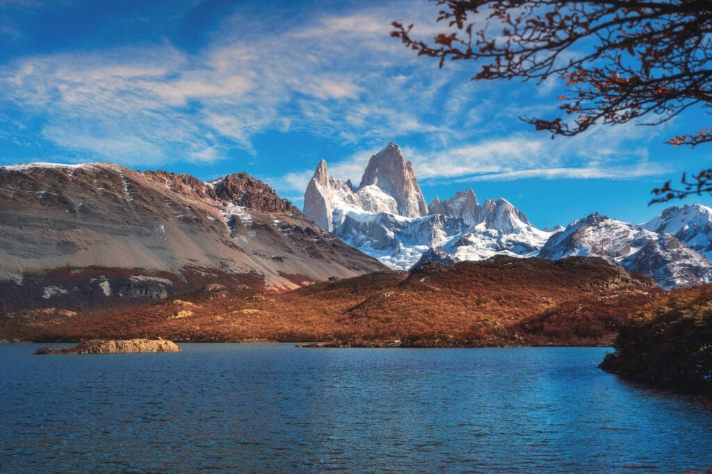 Majestic Fitz Roy mountain, a snow-capped granite peak, rising above a deep blue lake and brown scrubland under a bright sky.