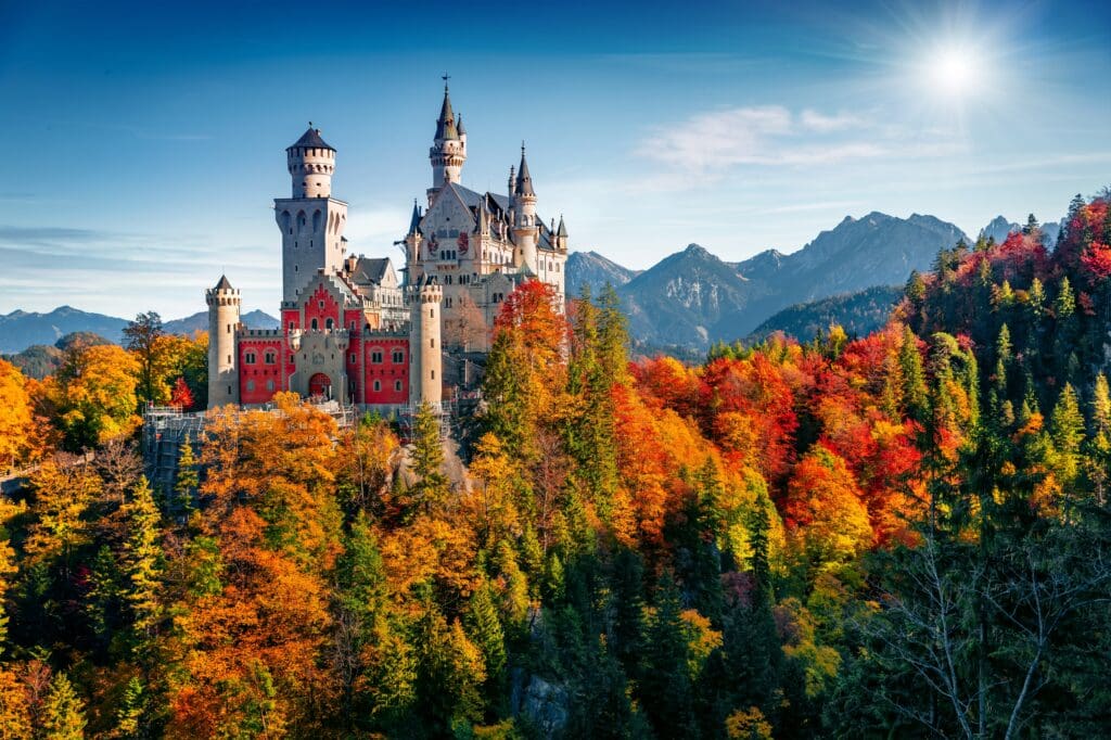 Neuschwanstein Castle, a 19th-century hilltop fortress, surrounded by a forest of fiery autumn colors under a bright sun and blue sky in the Bavarian Alps.