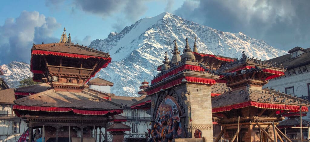 Historic, multi-tiered temples and structures with red trim in Patan Durbar Square, set against a backdrop of a massive snow-capped Himalayan mountain.