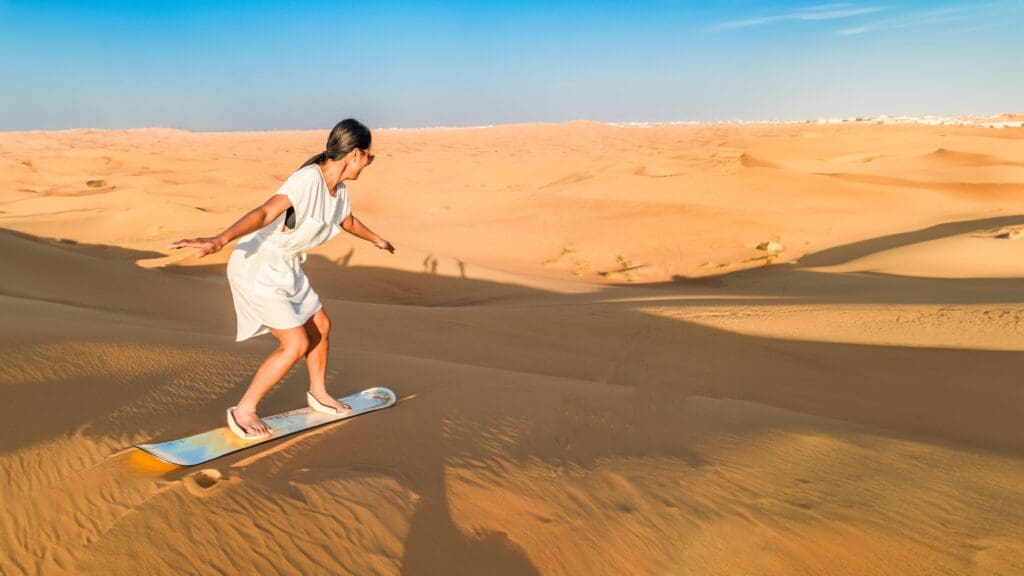 An Asian woman in a white dress is sandboarding down a large, sunlit desert dune in Dubai.