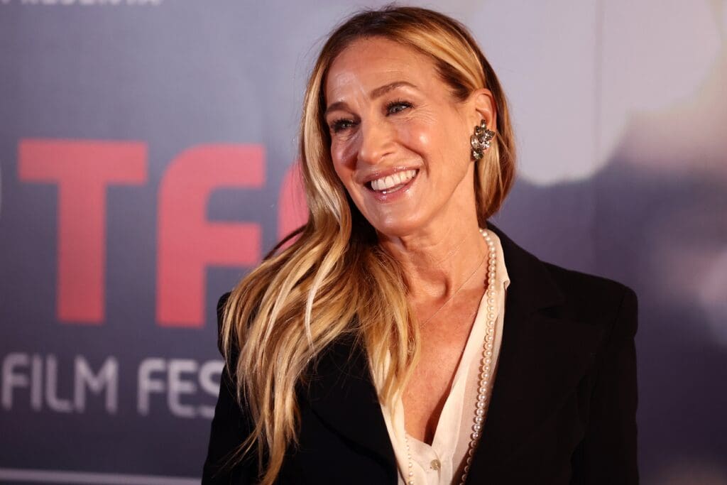 Actress Sarah Jessica Parker smiling broadly on the red carpet, wearing a black blazer, pearl necklace, and large earring.
