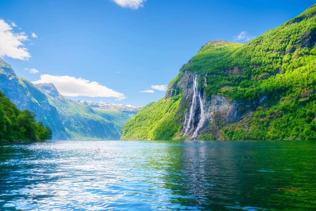 The Seven Sisters Waterfall cascades down a lush green cliff face into the bright blue waters of Geiranger Fjord, Norway, under a clear sky.