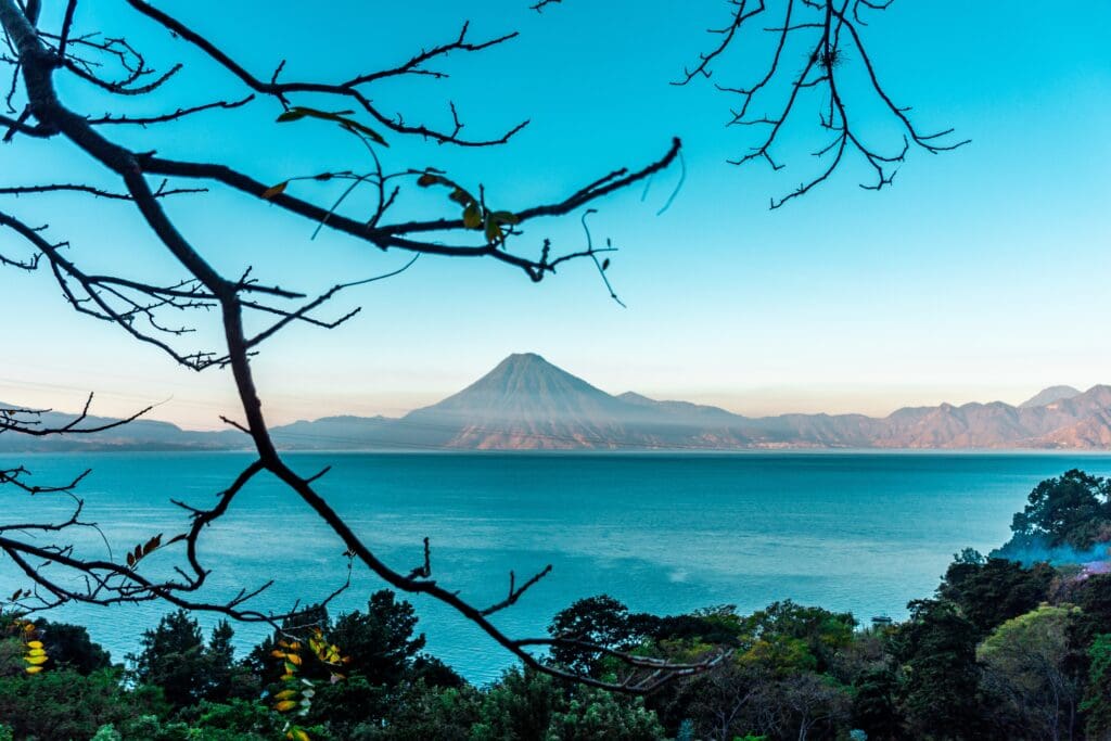 A volcano rises above the blue Lake Atitlán at sunset, framed by tree branches.