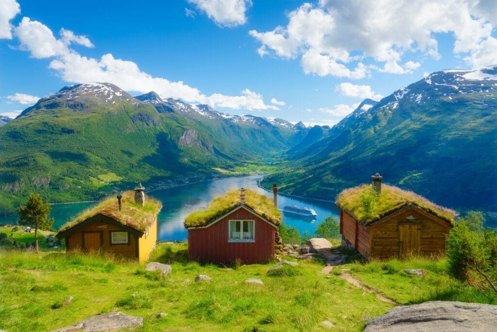Traditional Norwegian wooden houses with grass roofs overlook a deep blue fjord with a cruise ship, surrounded by green, steep mountains and snow-capped peaks under a bright blue sky.