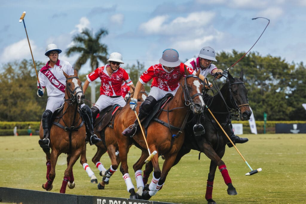 Four polo players on galloping horses aggressively chase the ball across a green field during a high-speed match.