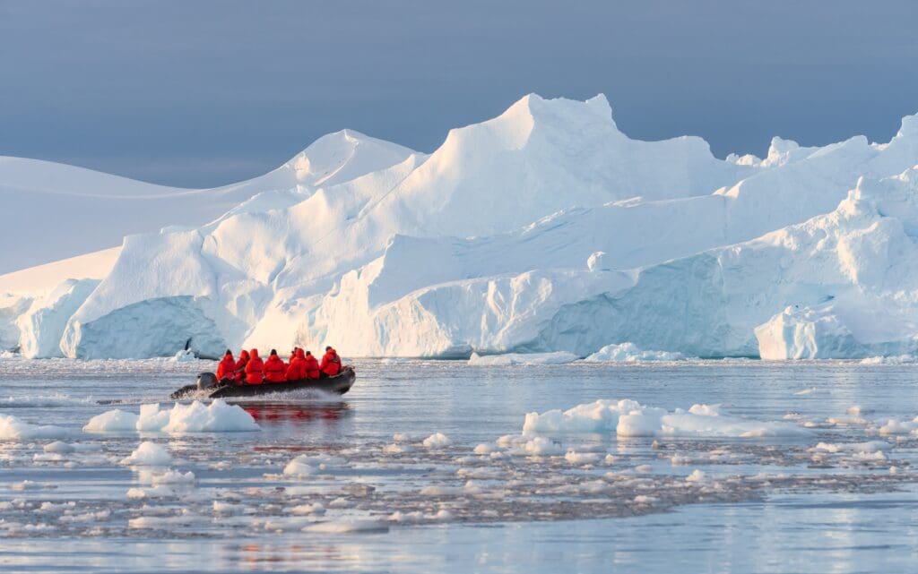 Cruise passengers in bright red parkas riding a black inflatable Zodiac boat near enormous blue and white icebergs in Antarctica