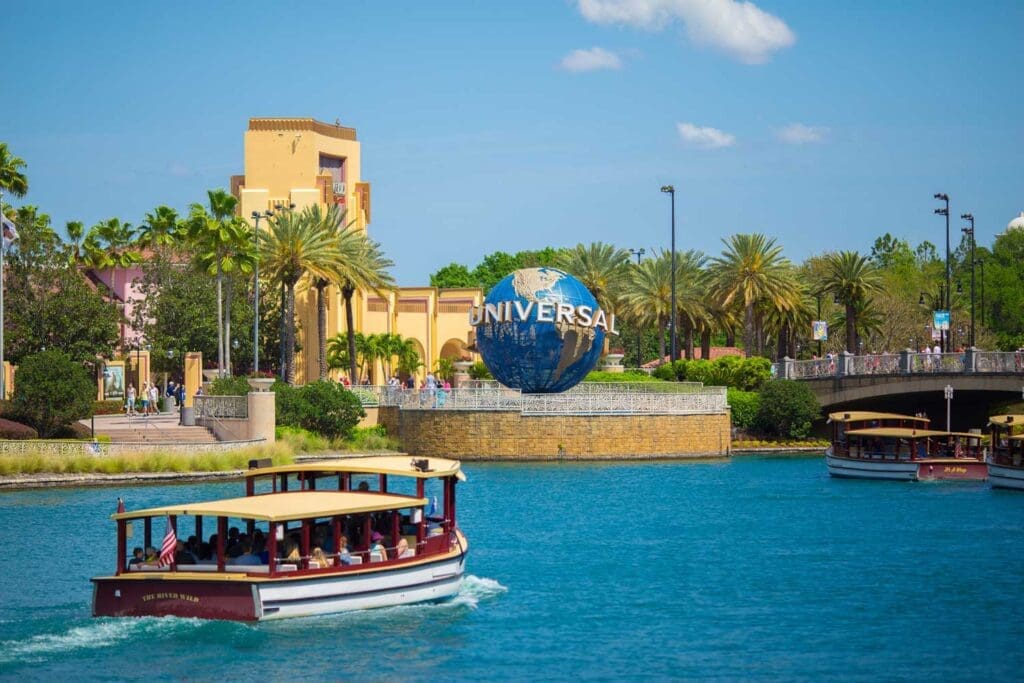 The Universal Studios globe in Orlando is seen from a boat on the water, with palm trees and a building in the background.